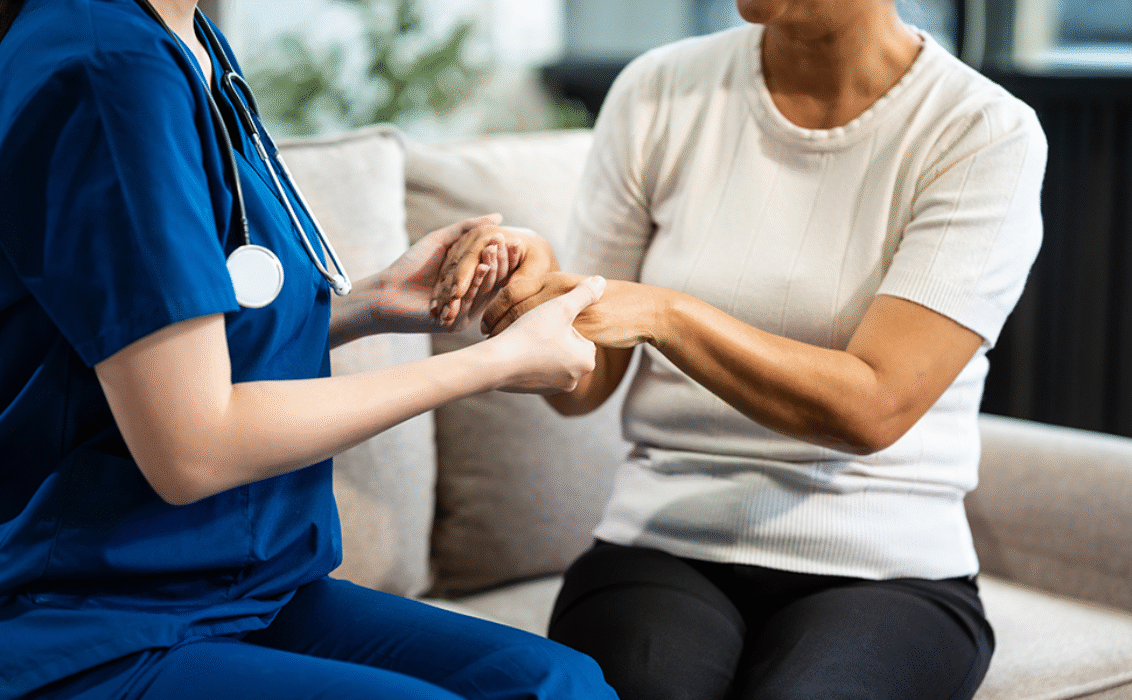 Una joven enfermera asiática, única mujer presente, coge de la mano a una mujer madura de pelo gris sentada en un sofá, ofreciéndole ánimos mientras realiza su historial médico y lleva a cabo controles de salud.